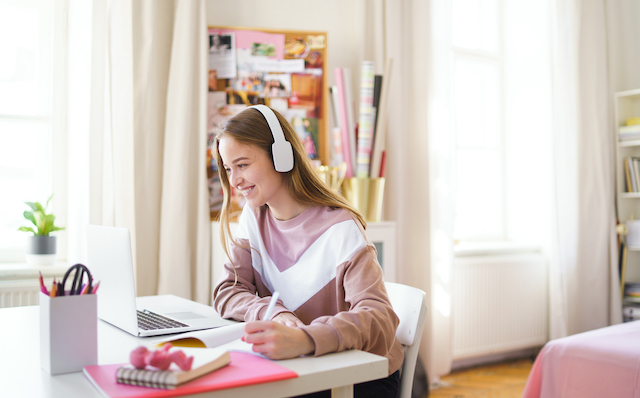 Young woman sitting at a desk looking at her laptop in a bedroom. 