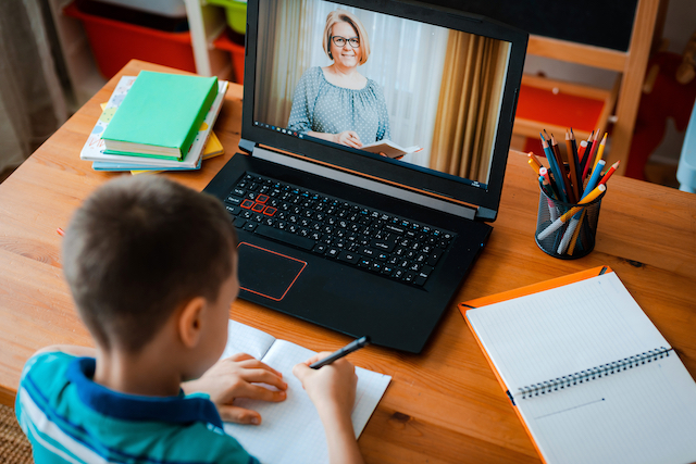 Elementary student taking notes and watching his teacher on an open laptop screen. 